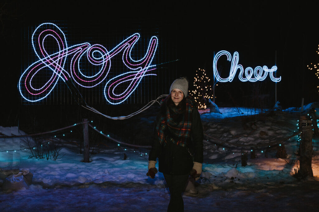 Woman standing in front of Christmas lights that say Joy and Cheer